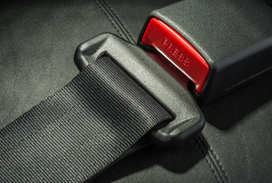 Close view of a seat belt assembly: woven webbing passing through a moulded latch plate next to a black buckle receiver with a red release button.