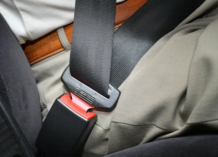A fastened seat belt running diagonally across a seated occupant, with the latch plate engaged in the buckle beside the seat.